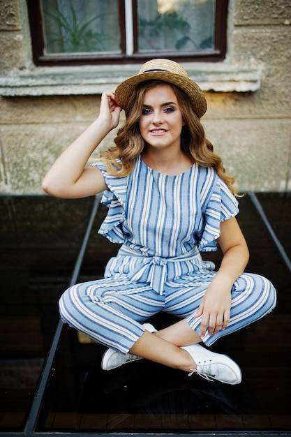 Free photo portrait of a fabulous young woman wearing striped overall and hat sitting on black shiny surface next to the building