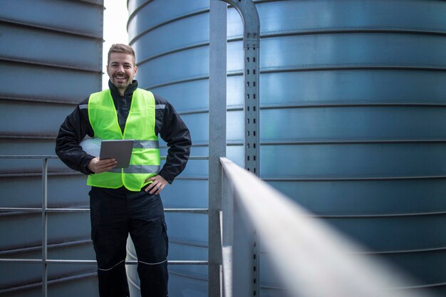 Portrait of factory engineer worker standing on metal platform between industrial storage tanks and looking to the camera