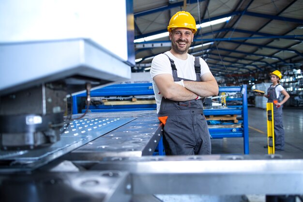 Portrait of factory worker in protective uniform and hardhat standing by industrial machine at production line