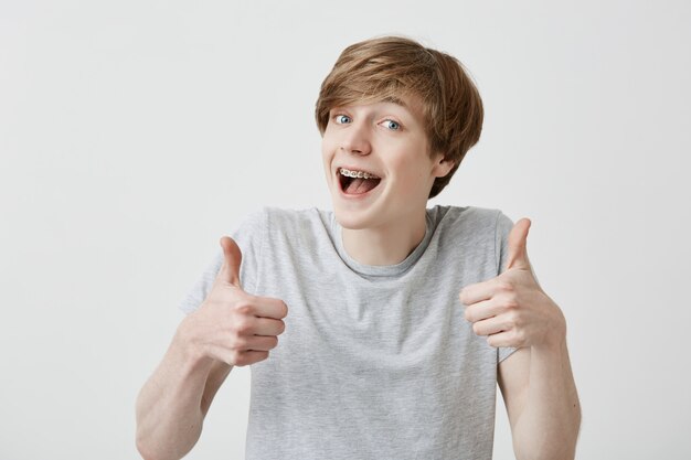 Portrait of fair-haired male student or customer with broad smile, looking at the camera with happy expression, showing thumbs-up with both hands, achieving study goals. Body language