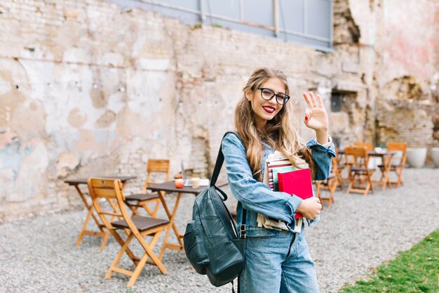 Portrait of fair-haired pretty female student carrying books with outdoor cafe on the background. Beautiful blonde girl in glasses showing OK hand sign and holding notes.