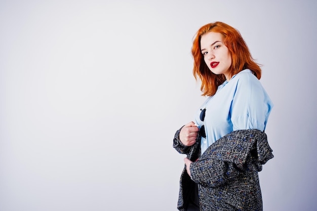 Portrait of a fantastic redheaded girl in blue shirt grey overcoat posing with sunglasses in the studio