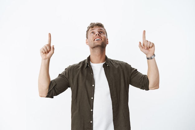 Portrait of fascinated and charmed amazed attractive blond guy with bristle smiling delighted raising head as looking impressed and happy up, pointing upwards over grey wall