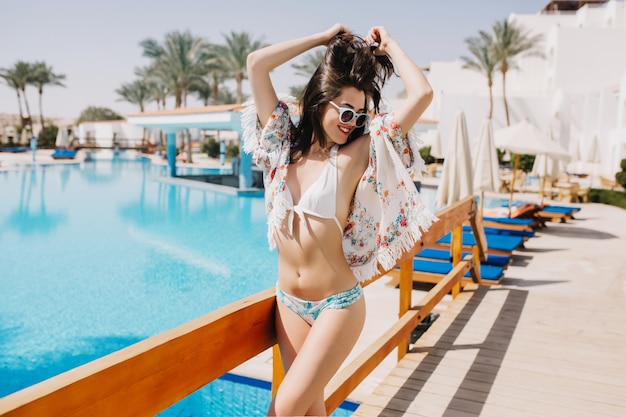 Portrait of fascinating cheerful girl in stylish sunglasses enjoys life at the resort. Slim brunette young woman in bikini posing next to blue pool, ready to have fun in summer vacation