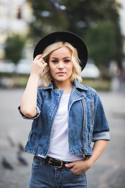Portrait of fashion blonde girl in city centre dressed up in jeans suite and white t-shirt and black hat