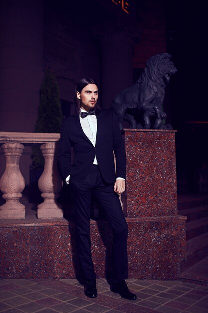 Portrait of fashion elegant long-haired young man. Attractive and handsome male model in black suit with mustache in the street at night