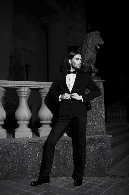 Portrait of fashion elegant long-haired young man. Attractive and handsome male model in black suit with mustache in the street at night