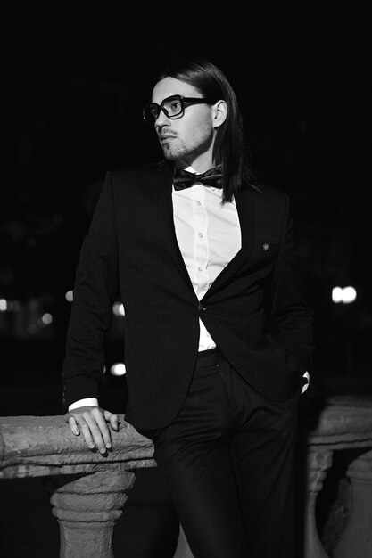 ファッションエレガントな長い髪の若い男の肖像画。夜の通りに口ひげと黒のスーツで魅力的でハンサムな男性モデル