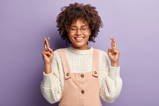 Free photo portrait of fashionable afro american woman keeps eyes shut with pleasure, smiles broadly, has fingers crossed, believes in good luck, asks for something, isolated over purple wall. hand gesture