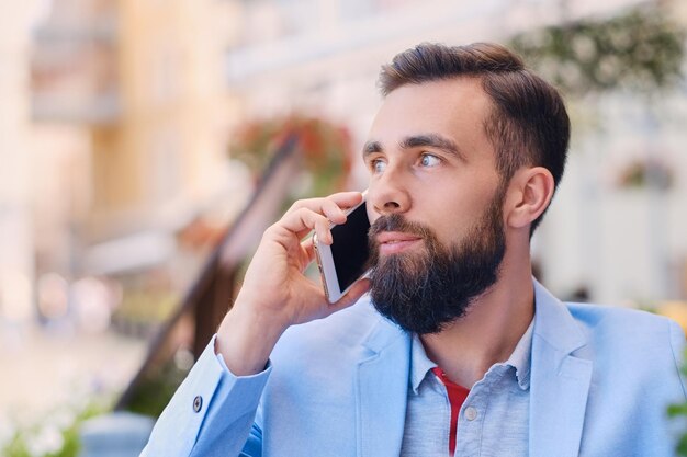 Портрет модного бородатого мужчины в синей куртке разговаривает по смартфону.