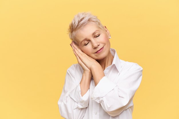 Portrait of fashionable blonde female on retirement posing isolated bending head, holding palms under cheek and keeping eyes closed, sleeping, napping, smiling with pleasure, having good dream