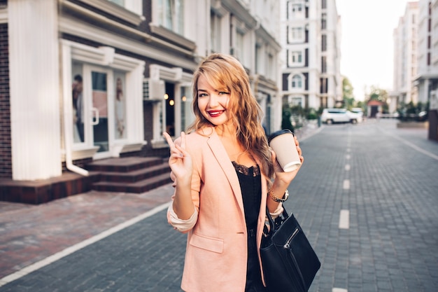 Portrait fashionable blonde woman with long hair walking in coral jacket on street. She holds a cup of coffee