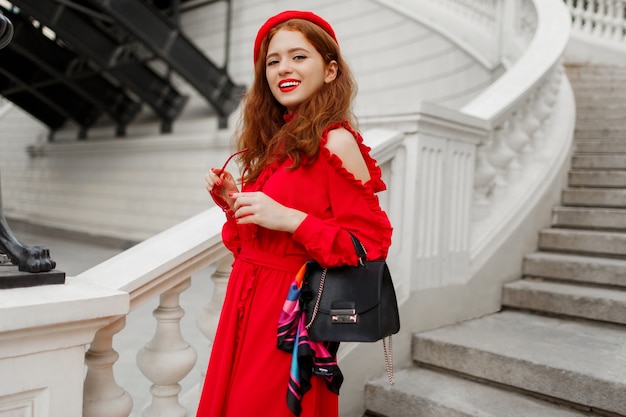 Portrait of fashionable ginger female in red beret and elegant dress posing outdoor.