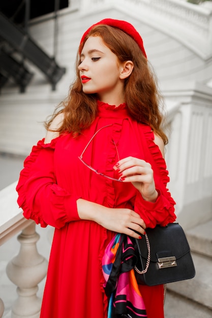 Portrait of fashionable ginger female in red beret and elegant dress posing outdoor.