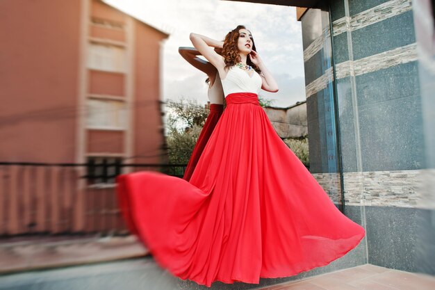 赤いイブニングドレスでファッショナブルな女の子の肖像画は、モダンな建物の背景ミラーウィンドウを提起空中のドレスを吹く