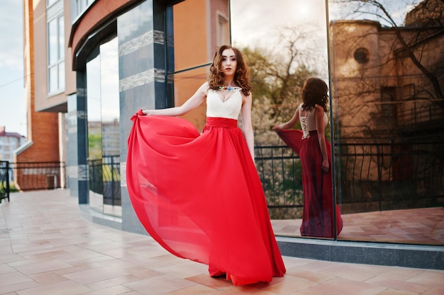 赤いイブニングドレスでファッショナブルな女の子の肖像画は、モダンな建物の背景ミラーウィンドウを提起
