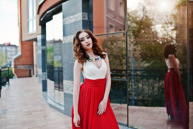 Portrait of fashionable girl at red evening dress posed background mirror window of modern building