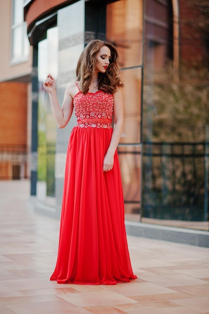 Portrait of fashionable girl at red evening dress posed background mirror window of modern building