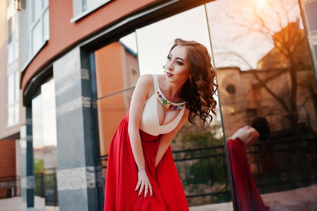 Portrait of fashionable girl at red evening dress with open bust posed background mirror window of modern building