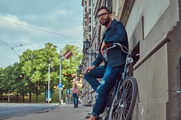 Portrait of a fashionable man in stylish clothes leaning against a wall with city bicycle on the street.