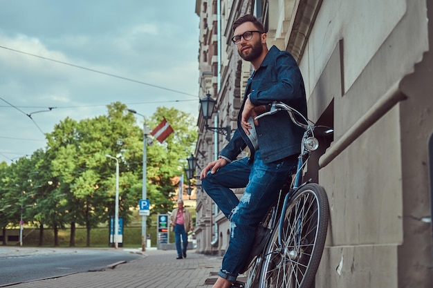 Portrait of a fashionable man in stylish clothes leaning against a wall with city bicycle on the street.