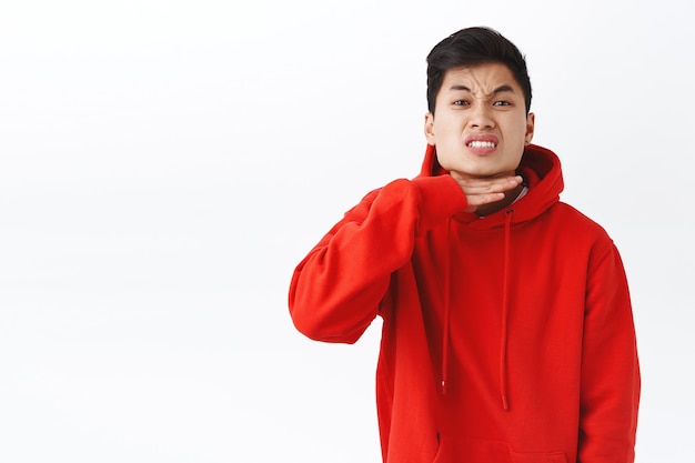 Portrait of fed up, pissed-off asian guy being angry, annoyed with someone, showing cut throat gesture as if head enough of bullshit, sick and tired of person telling lies, stand white wall.