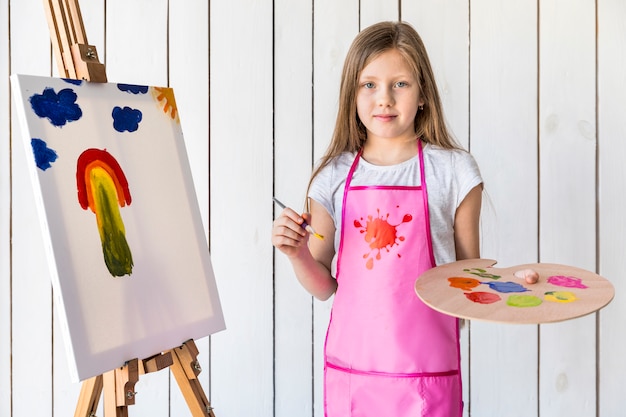 Portrait of a female artist holding paint brush and palette standing against white wooden wall