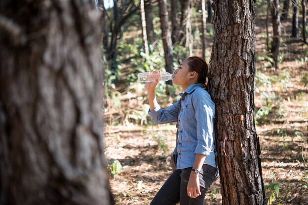 Portrait of female backpacker drinks fresh water from the bottle while carrying backpack in the pine forest.