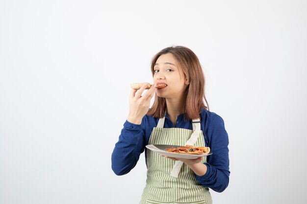 Portrait of female cook in apron eating slice of pizza on white 