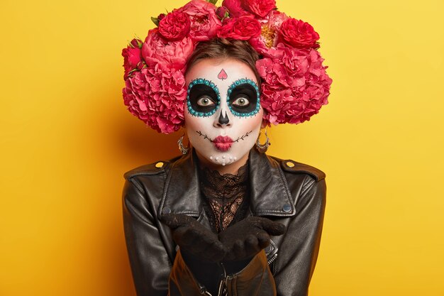 Портрет женского щеголеватого скелета готовится к мексиканскому карнавалу, дует воздушный поцелуй, носит макияж черепа, одет в черную куртку, имеет пугающий вид, изолированный на желтом фоне. Бодиарт и фейс-арт
