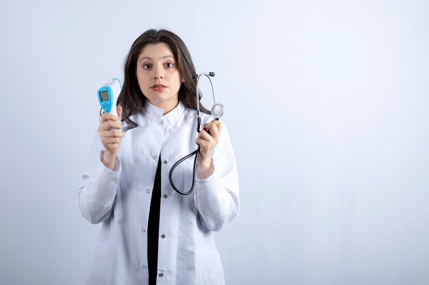 Portrait of female doctor holding thermometer and stethoscope on white wall.
