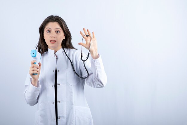 Portrait of female doctor showing thermometer and stethoscope on grey. 