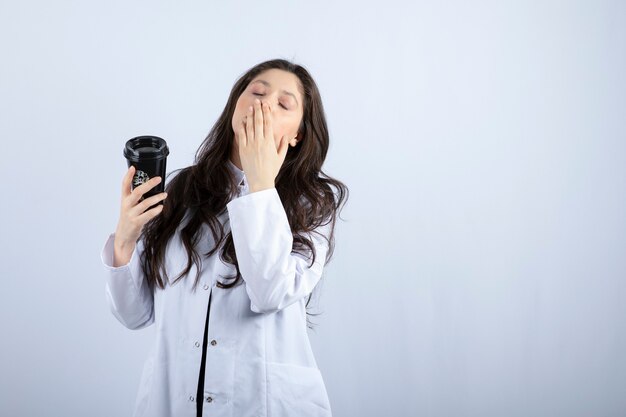 Portrait of female doctor with cup of coffee sleeping on white wall.