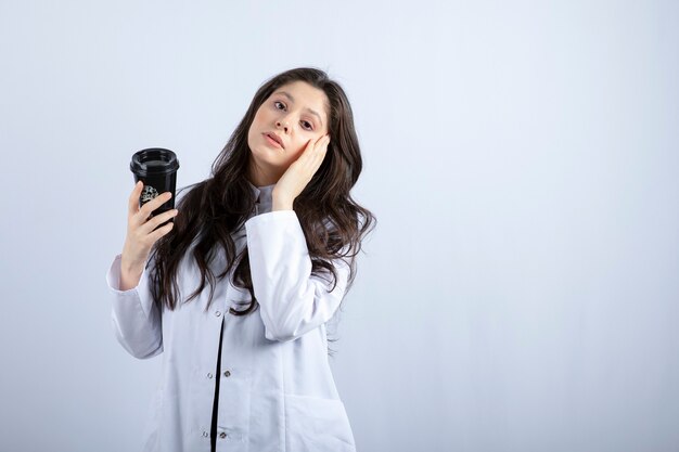 Portrait of female doctor with cup of coffee standing on grey. 