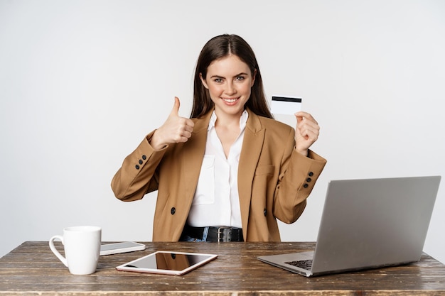 Portrait of female entrepreneur sitting at office table, showing credit card and thumbs up, recommending, standing over white background
