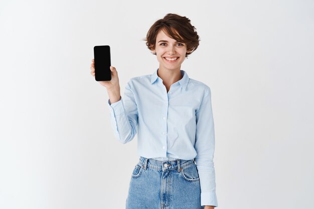Portrait of female entrepreneur smiling and showing empty smartphone screen, standing in blue collar shirt on white wall