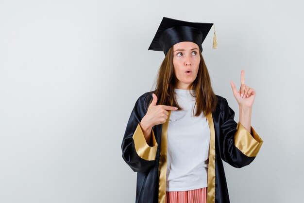 Portrait of female graduate pointing up and right in academic dress and looking hesitant front view