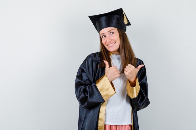 Portrait of female graduate showing double thumbs up in uniform, casual clothes and looking happy front view