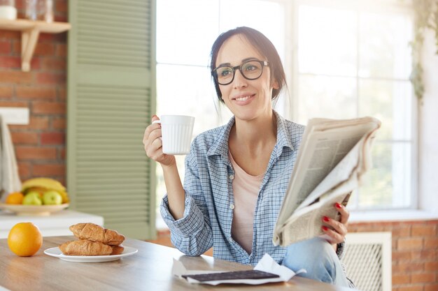 Portrait of female has breakfast, drinks coffee or tea with croissants and chocolate, reads newspaper alone at home.