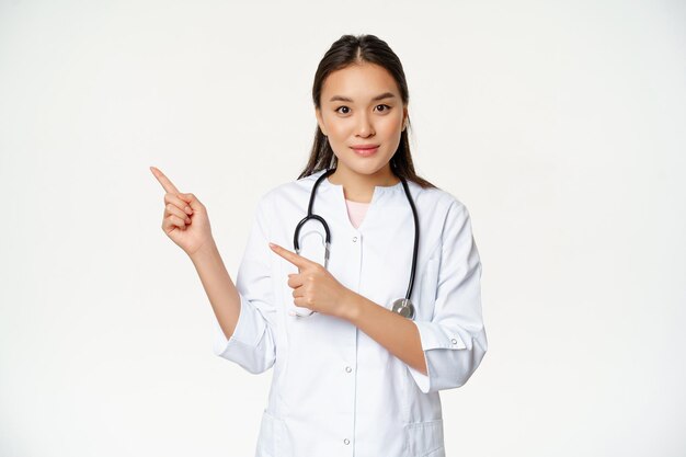 Portrait of female nurse in medical uniform, pointing fingers left, showing hospital related content, healthcare information aside, white background