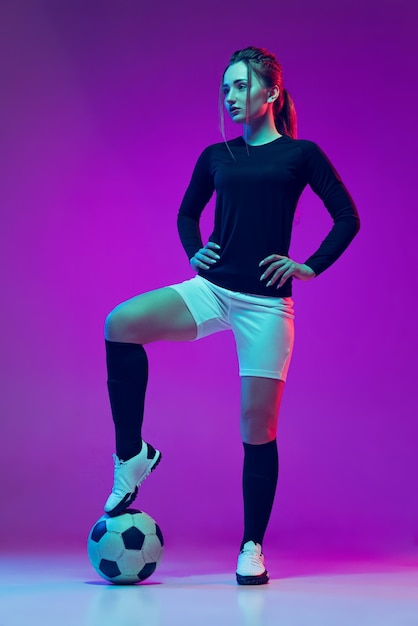 Portrait of female professional football player posing in uniform with ball isolated over purple background in neon