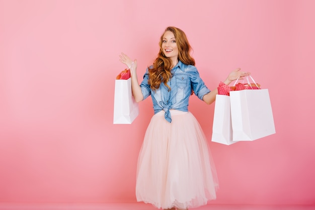 Портрет женского шопоголика, держащего бумажные пакеты из любимых магазинов и улыбающегося, изолированного на розовом фоне. Привлекательная молодая женщина с вьющимися волосами возвращается из покупок с пакетами
