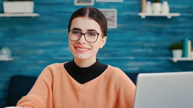 Portrait of female student using smartphone to browse internet and smiling at camera in living room. Young adult doing research work for school project assignment, working on mobile phone.