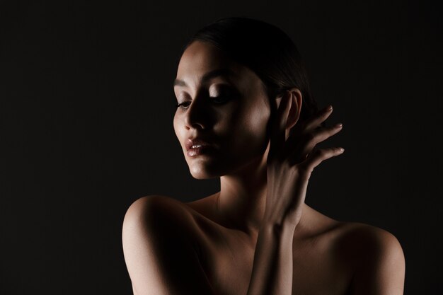 Portrait of feminine tender woman with sensual look looking aside in low lights, isolated over black