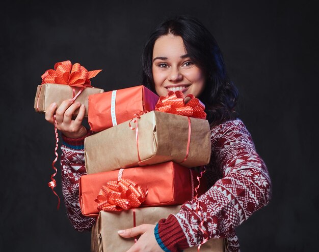 Portrait of festive smiling brunette female with long curly hair, dressed in a red sweater holds Christmas gifts.