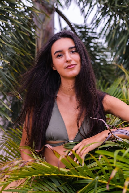 Portrait of fit tanned slim woman in green tiny bikini posing with tropical leaves