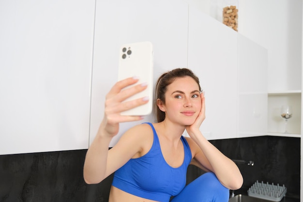 Free photo portrait of fitness girl posing for photo taking selfie on smartphone app sitting in kitchen wearing