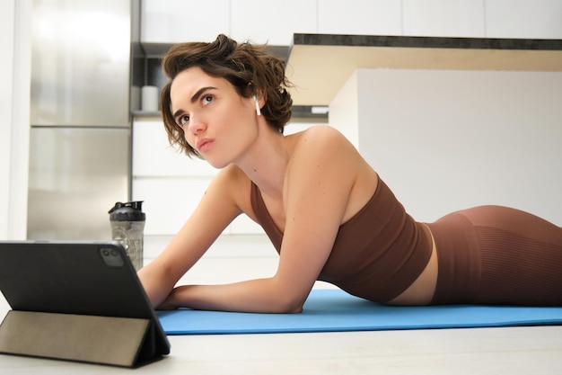 Free photo portrait of fitness girl with thoughtful face lying on rubber yoga mat at home listening and watchin