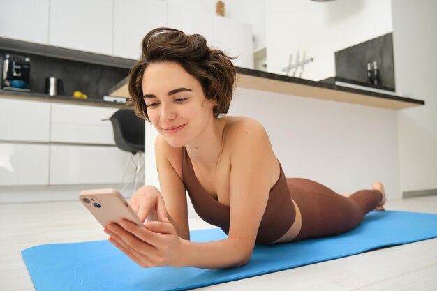 운동하는 동안 휴대폰을 사용하여 스마트폰을 보고 요가 매트에 누워 있는 피트니스 여성의 초상화