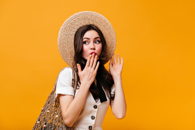 Portrait of flirtatious woman in straw hat and white T-shirt covering her mouth with hand and holding shopping bag on orange background.